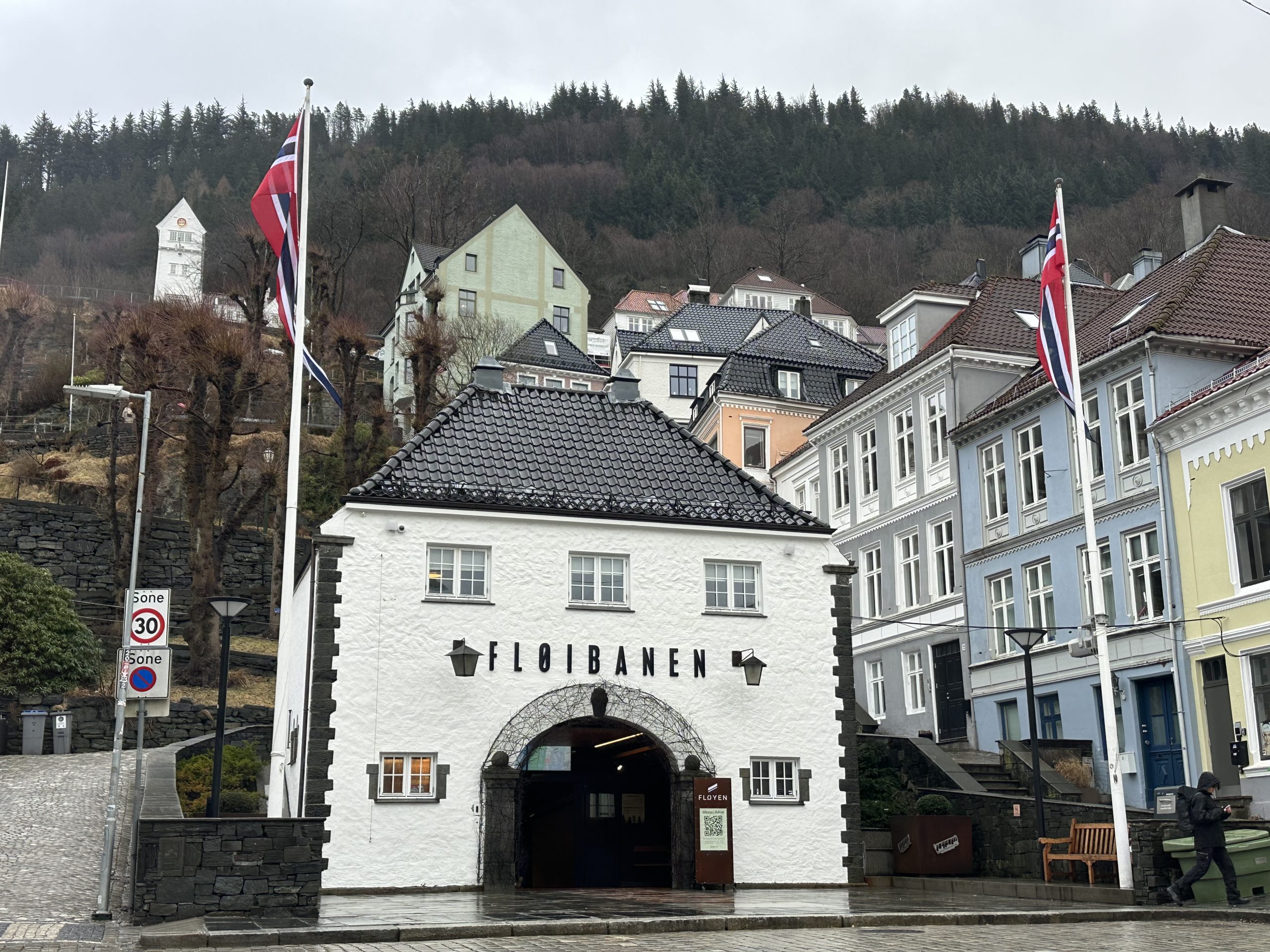 Estación del funicular Floibanen en Bergen, un edificio blanco con banderas de Noruega al pie de una colina arbolada