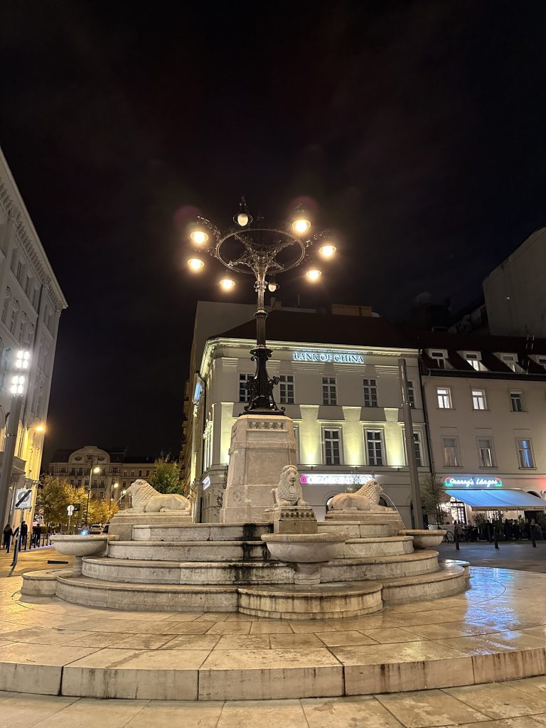 Plaza Vörösmarty de Budapest iluminada de noche con fuente y edificios históricos