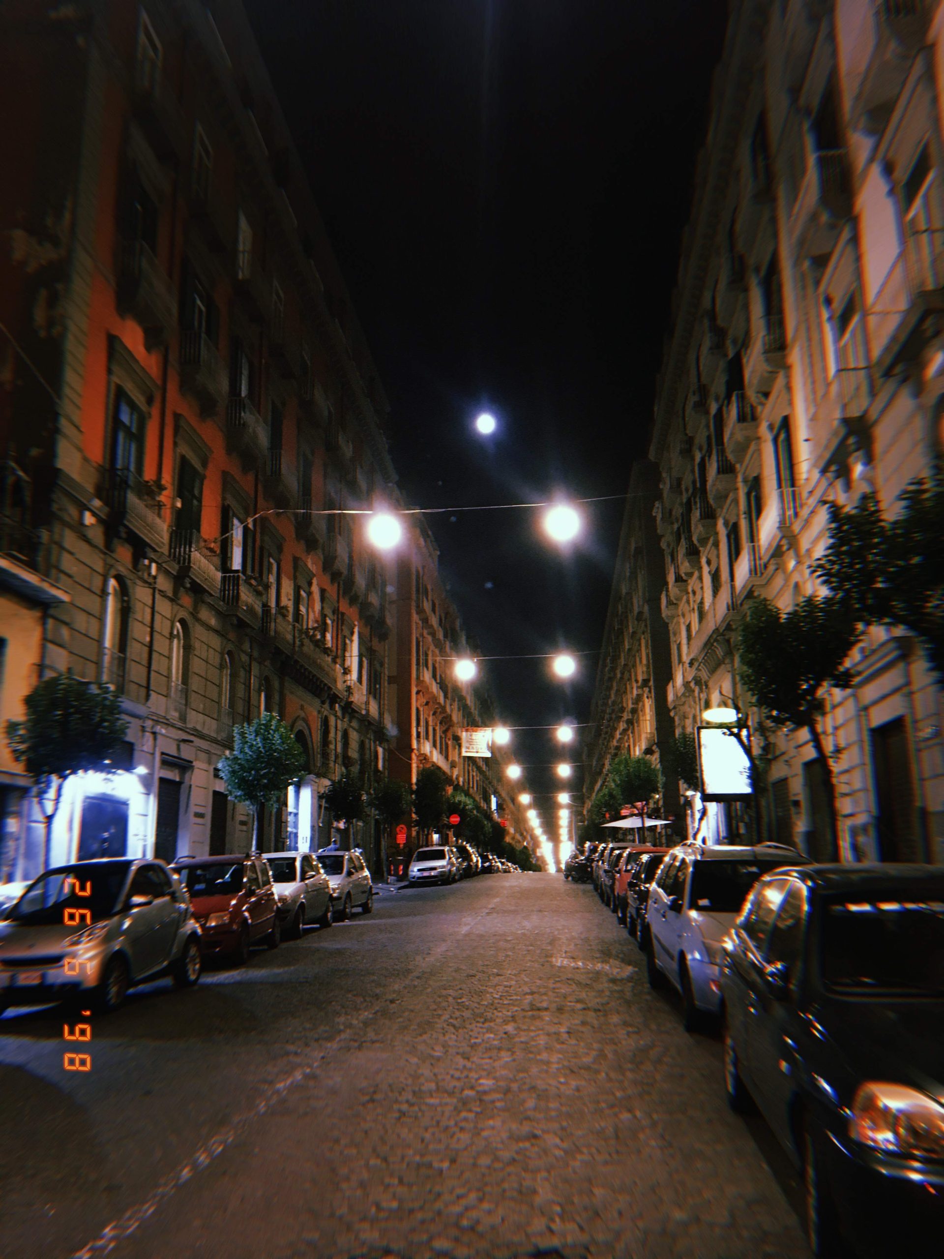 Calle del centro de Nápoles iluminada de noche con edificios históricos y coches aparcados.