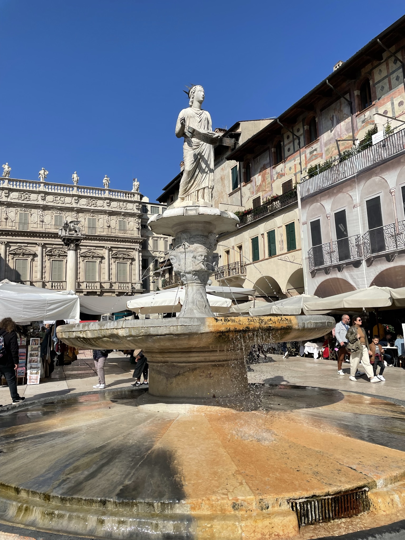 Fontana Madonna Verona en la Piazza delle Erbe, con edificios históricos y terrazas en el centro de Verona.